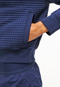 Suéter de punto texturizado azul marino con un patrón acolchado, que cuenta con bolsillos laterales y puños ribeteados. La tela parece suave con un ajuste relajado.