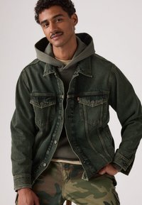 Σκούρο πράσινο τζιν μπουφάν με δύο μπροστινές τσέπες, κουμπιά κλεισίματος και ελαφρώς ξεθωριασμένη υφή. Φορεμένο πάνω από γκρι φούτερ με κουκούλα, συνδυασμένο με παντελόνια καμουφλάζ.