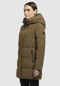 Brun pufferjakke med hætte, som har en frontlynlås, elastisk trim og to lynlåslommer på siden. Glat, isoleret materiale.