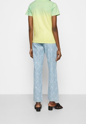 Ombre t-shirt z krótkim rękawem przechodzące od zieleni do żółci, w połączeniu z jasnoniebieskimi spodniami ozdobionymi wzorem z logo na całej powierzchni. Model ma na sobie czarne sandały.