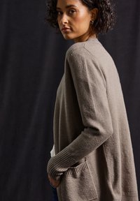 Cardigan tricoté beige clair avec une ouverture frontale, des poignets côtelés et des poches latérales. Tissu texturé, manches longues et coupe décontractée.