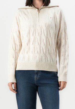 Kvinde iført cremefarvet kabelstrikket sweater med halv lynlåskrave og blå jeans, stående mod en hvid baggrund.