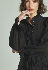 Blusa negra con detalles de volantes, mangas largas transparentes y cuello alto. Acentuada con botones decorativos a lo largo del plastrón delantero.