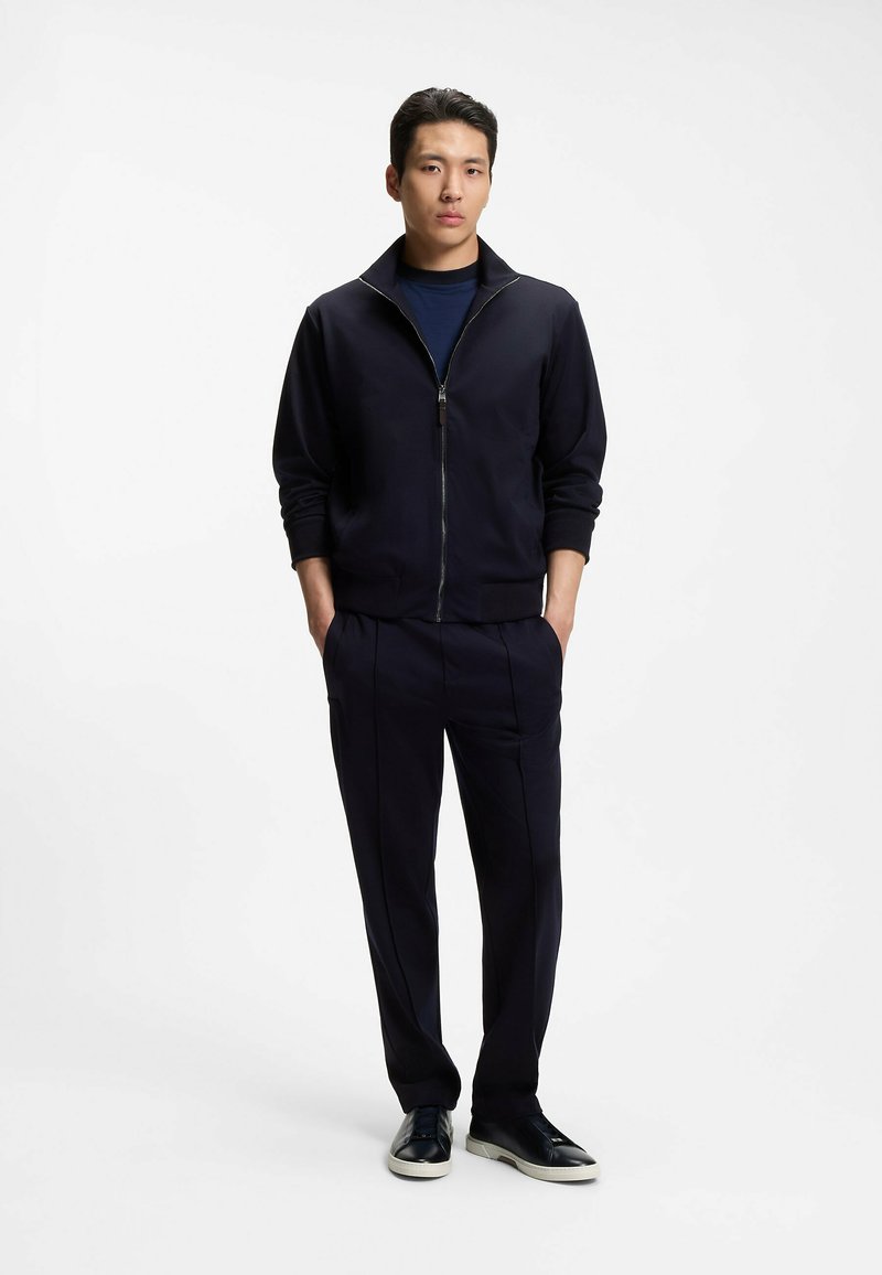 Homme debout les mains dans les poches, portant une veste noire zippée, un pantalon foncé et des baskets noires sur fond blanc.