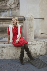 Crvena pamučna haljina s nabranim rukavima, bijeli džemper prebačen preko ramena, smeđe čizme od brušene kože do koljena, na pozadini od kaldrme.