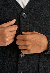 Tummanharmaa neuletakki, jossa on resorinen kaulus ja hihansuut sekä mustat napit. Kädet ovat kiinnittämässä nappeja valkoisen paidan päällä.