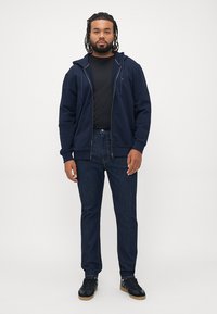 Homme avec des dreadlocks portant un hoodie zippé bleu marine, un t-shirt noir, un jean foncé et des baskets noires, se tenant devant un fond blanc uni.