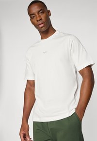 Hvid bomulds T-shirt med korte ærmer og rund halsudskæring, med et lille gråt logo på brystet. Kombineret med grønne bukser.