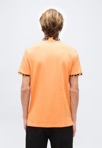 Homme portant un polo à manches courtes orange avec des accents floraux noirs sur le col et les bords des manches, vu de dos.