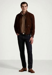 Hnedá semišová bunda so zipsom, béžové polo tričko, tmavé džínsy a hnedé kožené topánky. Model stojí na neutrálnom pozadí.