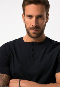 Navy blauw T-shirt met korte mouwen, voorzien van een ronde kraag, een placket met vier knopen en een zachte katoenen textuur. Zichtbare tatoeages op de onderarm.