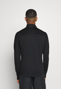 Maglietta sportiva nera a maniche lunghe con colletto alto, caratterizzata da una texture liscia e un design aderente. Stile minimalista senza loghi visibili.