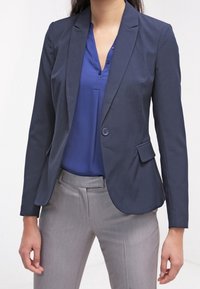 Granatowy blezer z pojedynczym guzikiem, wyciętymi klapami i przednią kieszenią, zestawiony z niebieską bluzką i jasnoszarymi spodniami. Gładka tkanina.