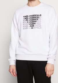 Sudadera blanca con el texto negro repetido "KARL LAGERFELD". Cuello redondo y puños acanalados que ofrecen un ajuste cómodo. Textura de tela suave y lisa.
