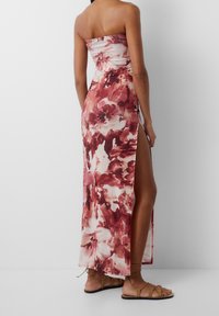 Robe longue sans bretelles avec un imprimé floral dans des teintes rouges et roses. Présente une fente sur le côté et est fabriquée dans un tissu fluide. Portée avec des sandales plates.