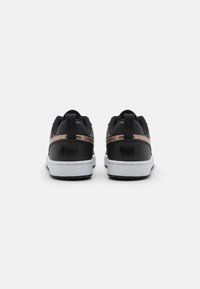 Nike sportcipő fekete felsőrésszel, bronz díszítéssel, fehér középtalppal és textúrált anyagból készült. A hátsó részen egy feltűnő logó található.