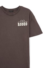 T-shirt in cotone marrone scuro con maniche corte, caratterizzato da una grafica "RODEO" color crema e motivi decorativi sul lato superiore sinistro.