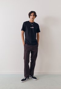 Sort t-shirt med hvidt logo, brune bukser og sorte sneakers med hvide såler. Modellen har bølget hår og en sølvhalskæde. Enkel baggrund.