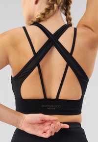Sutien sport negru cu bretele încrucișate și design cu spate decupat. Material neted, elastic, cu un logo pe bandă. Conceput pentru purtare activă.