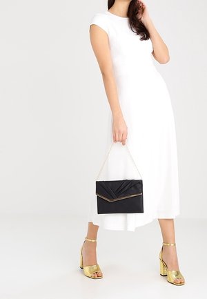 Černá psaníčko s plisovaným designem a zlatým lemováním, držené osobou v bílých šatech. Viditelné zlaté sandály na podpatku s pásky kolem kotníku.