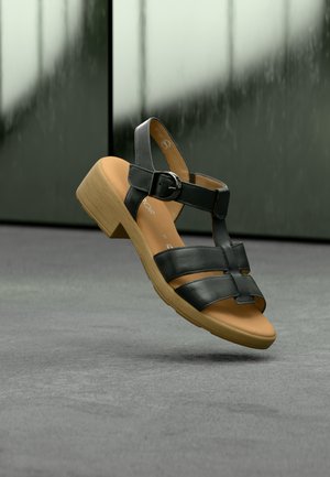 Μαύρο δερμάτινο σανδάλι με ανοιχτό δάχτυλο, ρυθμιζόμενο λουρί και χαμηλό ξύλινο τακούνι. Η επιφάνεια πάτου είναι καφέ με λεία υφή.