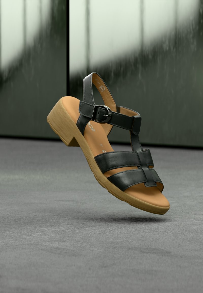 Μαύρο δερμάτινο σανδάλι με ανοιχτό δάχτυλο, ρυθμιζόμενο λουρί και χαμηλό ξύλινο τακούνι. Η επιφάνεια πάτου είναι καφέ με λεία υφή.