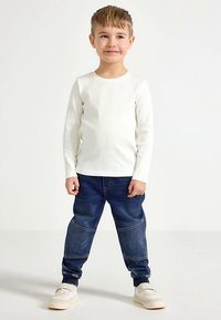 Maglietta bianca a maniche lunghe, pantaloni jogger di denim blu con pannelli sulle ginocchia e sneakers beige. Bambino in piedi con le mani rilassate ai lati, sorridente.