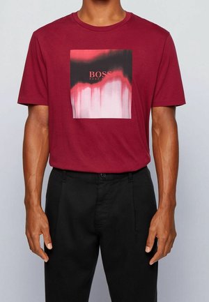 Man draagt een bordeauxrood t-shirt met een zwart, rood en wit abstract vierkant grafisch patroon en zwarte broek tegen een grijze achtergrond.