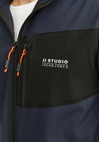 Jūros mėlynumo ir juodos spalvos zipseris su pliušiniu apykakle, kuriame yra oranžinis veržliarakčio vienetas ir baltas logotipas ant krūtinės kišenės.