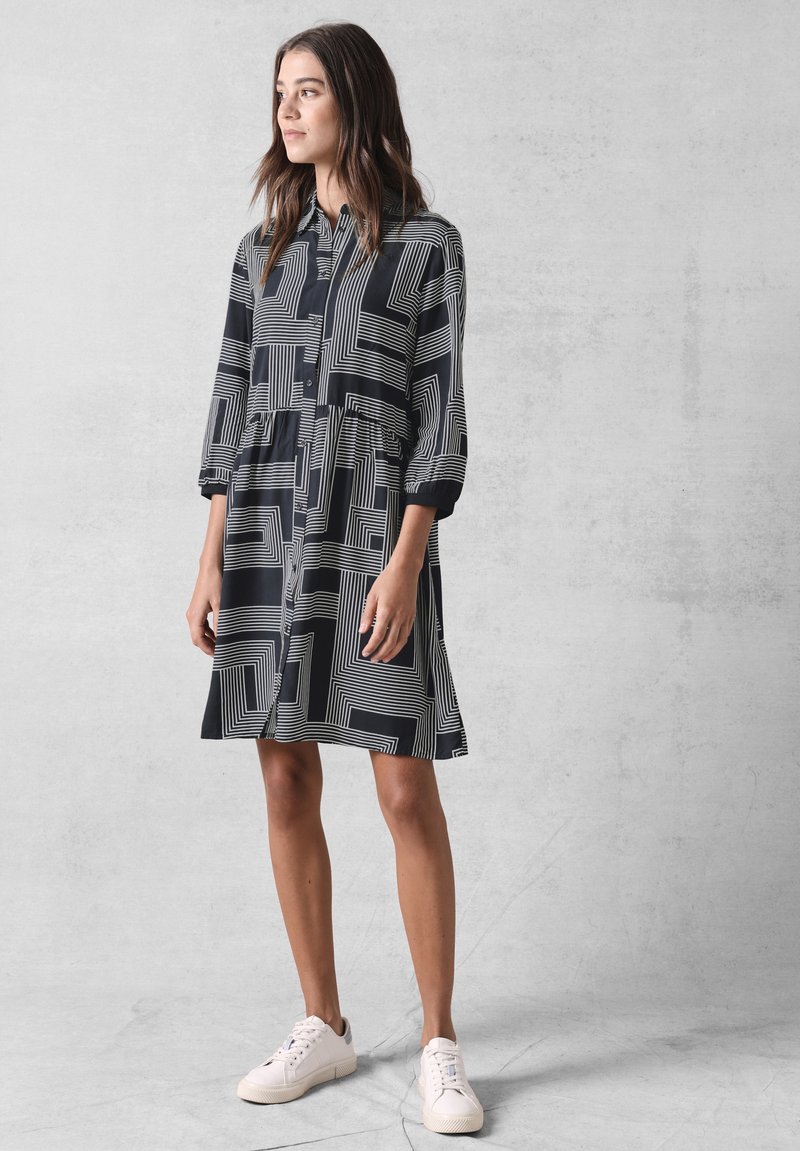 Abito button-down bianco e nero con motivi geometrici a linee, maniche a tre quarti e vestibilità comoda. Orlo svasato sopra il ginocchio.