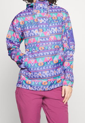 Persona que lleva una chaqueta con capucha de patrón geométrico multicolor y pantalones morados y rosas, de pie contra un fondo claro y liso.
