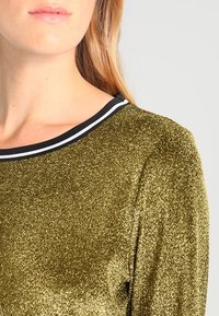 Eine blonde Person trägt ein glänzendes goldenes Oberteil mit einem schwarz-weißen gestreiften Halsausschnitt, von der Schulter bis zum Mund vor einem schlichten Hintergrund gezeigt.