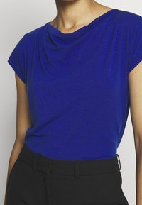 Top azul drapeado en una tela ligera con un ajuste relajado y mangas cortas, combinado con pantalones negros de tiro alto.