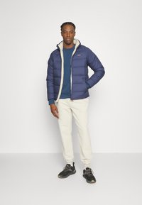 Veste matelassée bleu marine avec une doublure en polaire blanche, associée à un pull bleu et un pantalon de jogging crème. Chaussures de sport noires avec des accents gris.