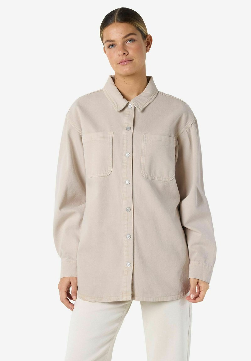 Világos bézs színű, gombos ing gallérral, hosszú ujjal, két mellzsebbel és puha, texturált anyaggal. Az elején ezüst színű gombok díszítik.