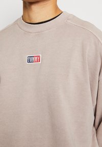 Sudadera beige con cuello redondo y puños acanalados, que presenta un pequeño logo bordado de "TOMMY" en azul, rojo y blanco. Textura suave.