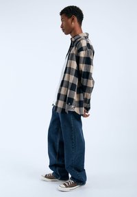 Camisa de franela a cuadros en beige y negro, de corte relajado, combinada con unos vaqueros azules de gran tamaño. El modelo lleva zapatillas marrones con suelas blancas.
