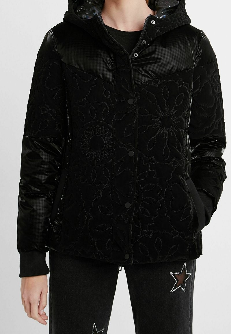 Femme portant une veste matelassée noire avec des motifs floraux en couture et des manches rembourrées brillantes, associée à un pantalon noir orné d'étoiles.