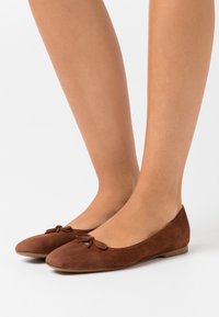 Ballerines en suede marron avec un bout arrondi et un petit nœud décoratif. Semelle plate et texture lisse. Convient pour un usage décontracté.