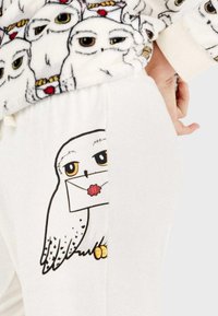 Pantalones de pijama blancos con un búho de dibujos animados sosteniendo una carta, detallados con contornos negros y pies amarillos, sobre una textura suave.