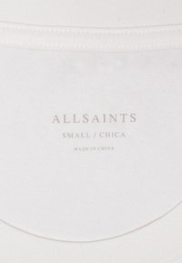 Hvidt stof med en glat tekstur, der har et mærke med teksten "ALLSAINTS, SMALL / CHICA, MADE IN CHINA" i lysegrå skrifttype.