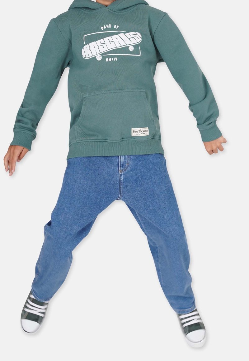 Grüne Kapuzenjacke mit Skateboardgrafik, mit einer vorderen Tasche, kombiniert mit blauen, weiten Jeans und grünen Turnschuhen mit weißen Akzenten.