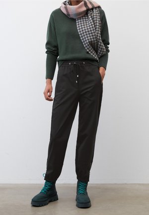 Pantalon noir avec taille à cordon, accents zippés sur le côté, associé à un pull vert et une écharpe à carreaux rose. Le modèle porte des bottes à lacets teal.