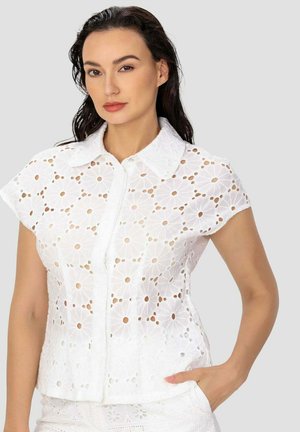 Femme aux cheveux foncés portant un chemisier blanc à manches courtes en broderie anglaise avec des motifs floraux découpés et un bas blanc, main dans la poche.