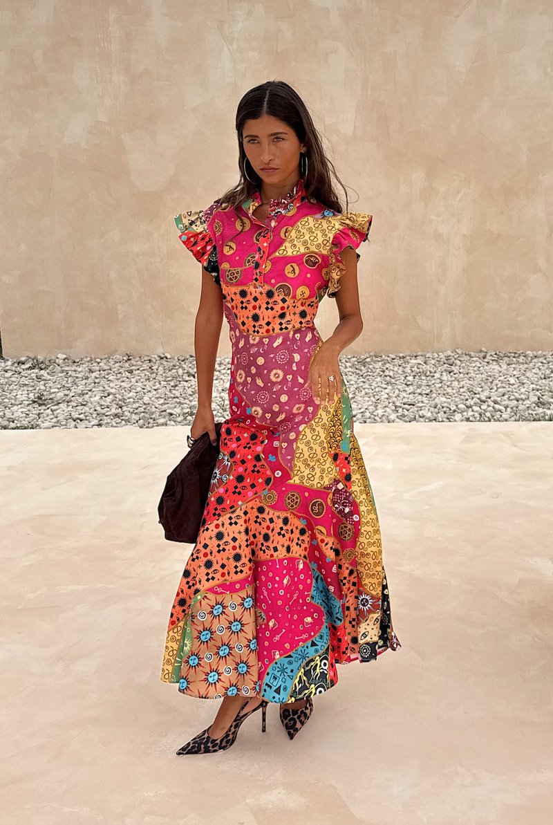 Kleurrijke patchwork jurk met gerimpelde mouwen, featuring diverse patronen in roze, geel, oranje en blauw. Model houdt een donkere handtas vast.