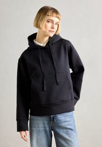 Felpa blu navy scuro realizzata in tessuto morbido, con tasca frontale, cordini e polsini a costine. Abbinata a jeans azzurri chiari.
