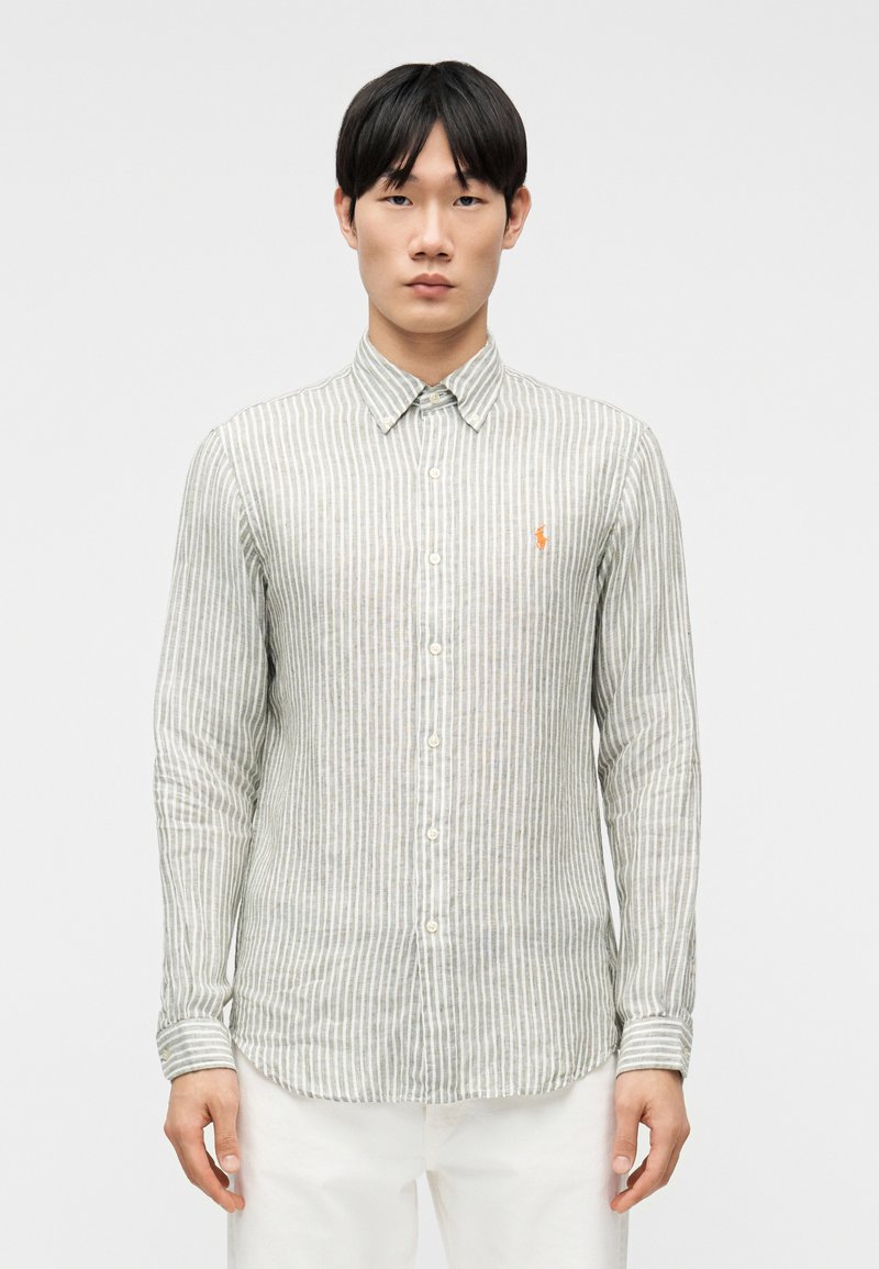 Νέος άνδρας όρθιος, κοιτάζοντας μπ ροστά, με ανοιχτό γκρι και λευκό πουκάμισο με κατακόρυφες ρίγες, κουμπωμένο, με πορτοκαλί λογότυπο και λευκό παντελόνι.