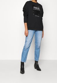 Sudadera negra con gráfico de lentejuelas "PARÍS", complementada con jeans de mezclilla azul claro y botas negras de tobillo. Presenta un corte relajado y un material texturizado.