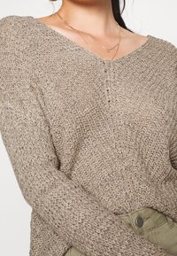 Beige stickad tröja med V-ringning, strukturerad tyg, långa ärmar och tre små knappar framtill, i kombination med olivgröna byxor.