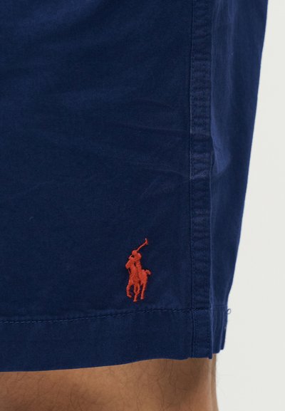 Σορτς σε ναυτικό μπλε χρώμα με κόκκινο κεντημένο λογότυπο παίκτη πόλο κοντά στο τελείωμα, φορεμένα στο πόδι ενός ατόμου με ορατό δέρμα.